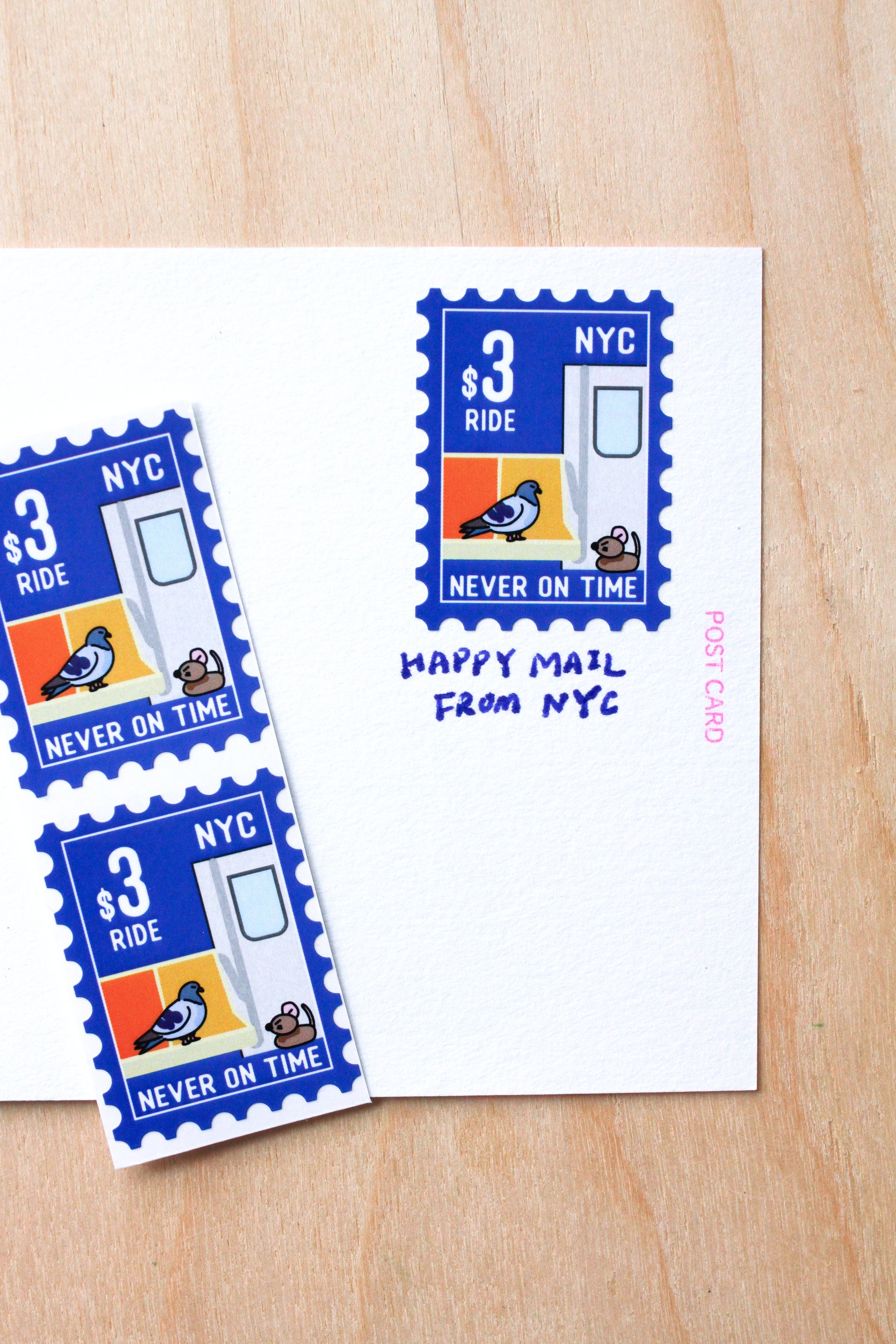 A postcard design shown on a light wood surface, featuring blue postage-stamp-style graphics with a bird and the words 'NYC $3 RIDE NEVER ON TIME,' plus a white postcard with the handwritten text 'HAPPY MAIL FROM NYC.'.