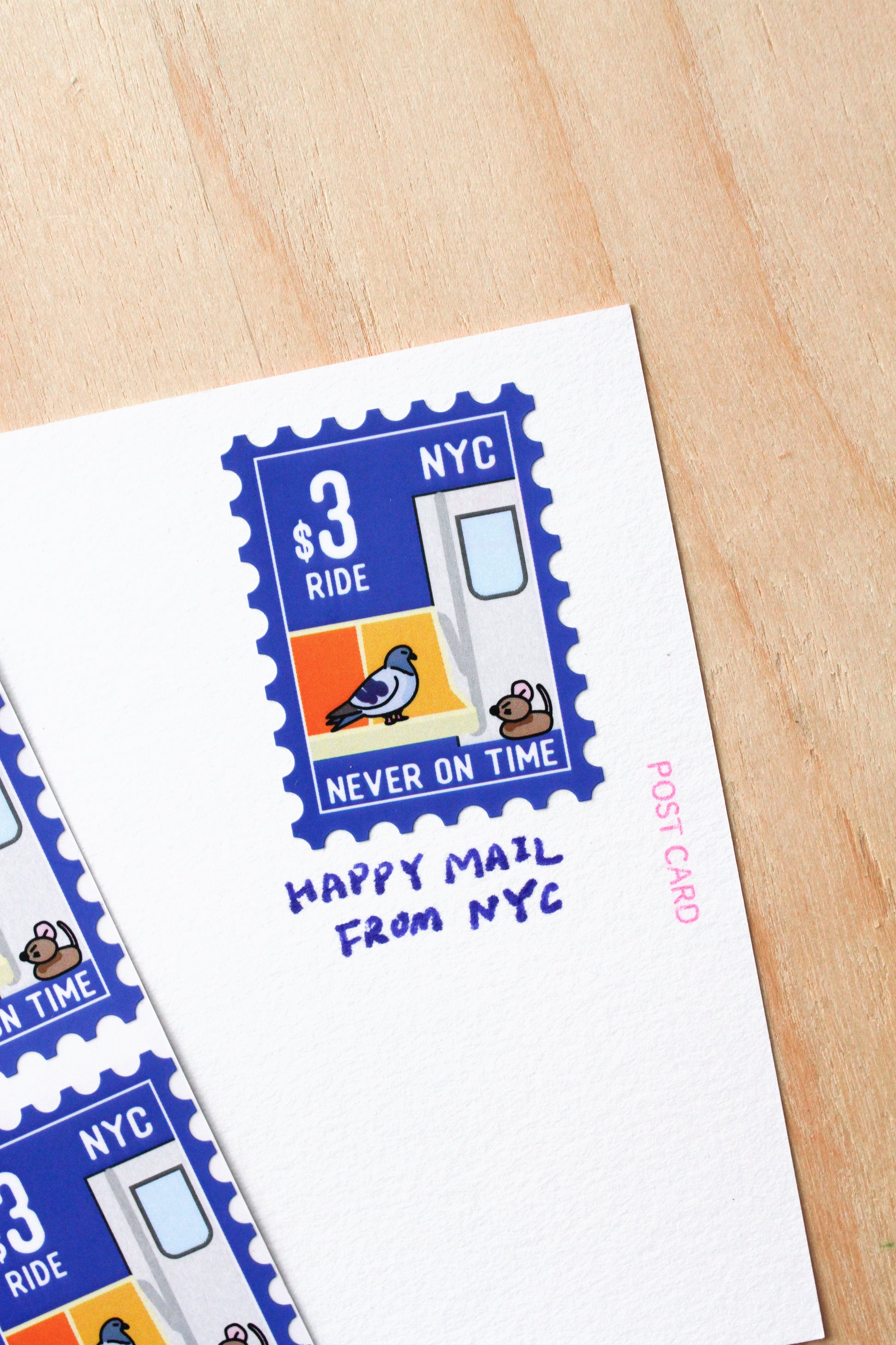 A white postcard printed with a blue postage-stamp-style NYC illustration and the text 'HAPPY MAIL FROM NYC', placed on a light wood surface with part of another postcard visible at the left edge.